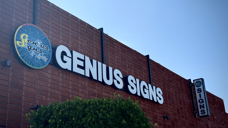 The Genius Signs & Lighting building: illuminated channel letters and a circular logo cabinet mounted on brick, with a projecting blade sign.