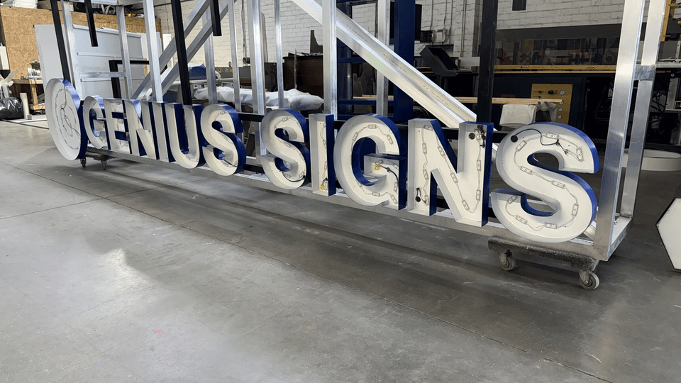 Channel letters reading GENIUS SIGNS part-built on a shop cart, LED modules and wiring still exposed, on the fabrication floor.